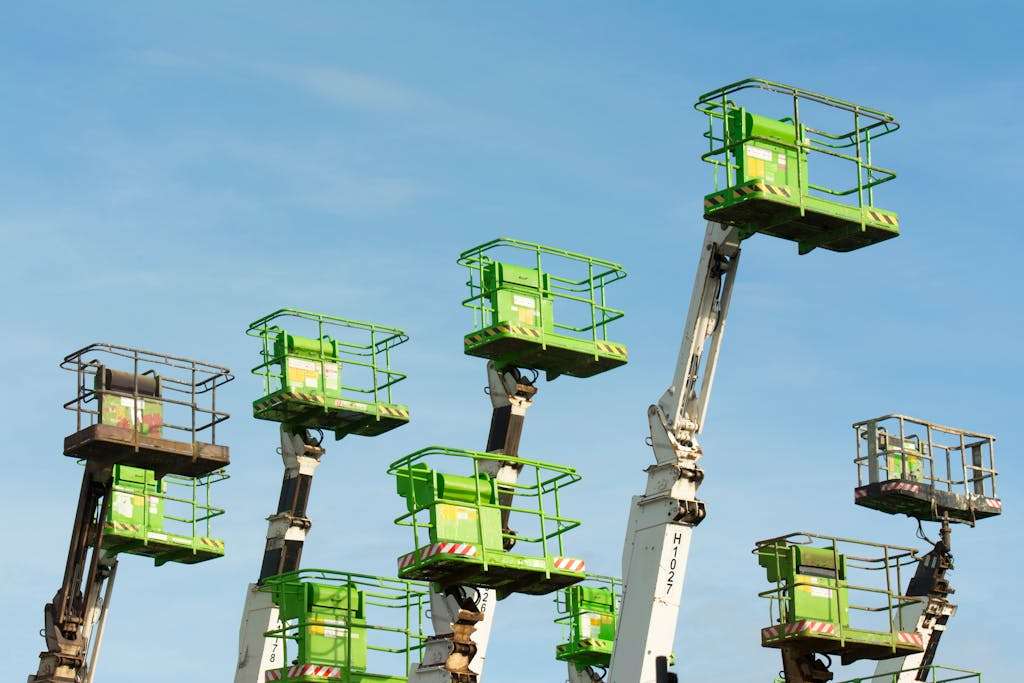 A group of green cherry pickers extending against a clear blue sky in an industrial setting.